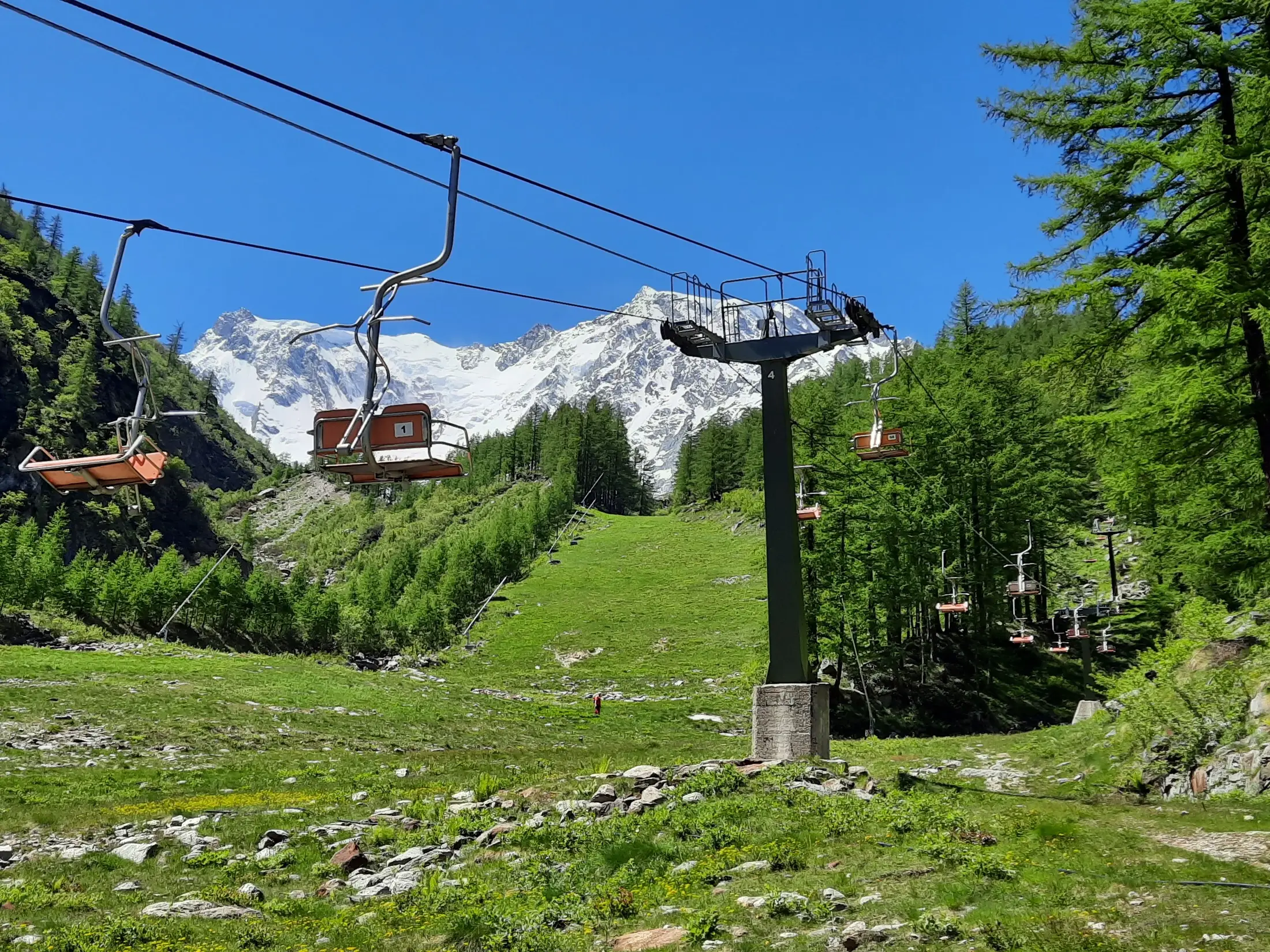 Panorama estivo dal Belvedere a Macugnaga, con sentieri di montagna, prati verdi e le vette del Monte Rosa sullo sfondo.